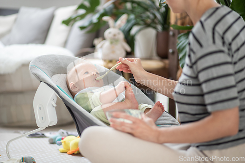 Image of Mother spoon feeding her baby boy infant child in baby chair with fruit puree. Baby solid food introduction concept.