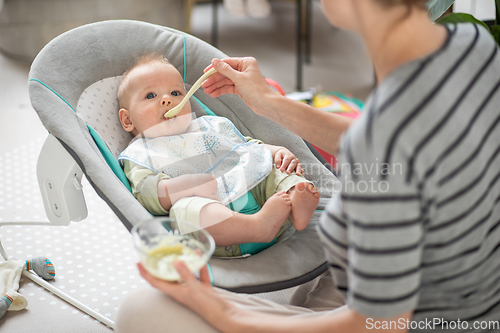 Image of Mother spoon feeding her baby boy infant child in baby chair with fruit puree. Baby solid food introduction concept.