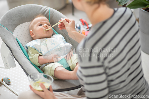 Image of Mother spoon feeding her baby boy infant child in baby chair with fruit puree. Baby solid food introduction concept.