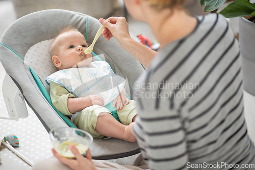 Image of Mother spoon feeding her baby boy infant child in baby chair with fruit puree. Baby solid food introduction concept.