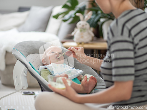 Image of Mother spoon feeding her baby boy infant child in baby chair with fruit puree. Baby solid food introduction concept.