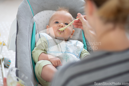 Image of Mother spoon feeding her baby boy infant child in baby chair with fruit puree. Baby solid food introduction concept.