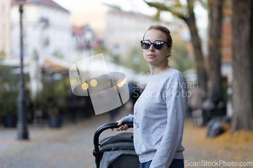 Image of Stylish young woman wearing sunglasses walking through Ljubljana city center pushing and rocking baby stroller. Warm autumn or spring weather for outdoor activity.