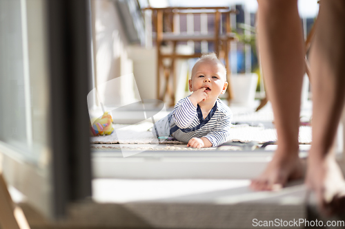 Image of Cute little infant baby boy playing with toys outdoors at the patio in summer being supervised by her mother seen in the background.