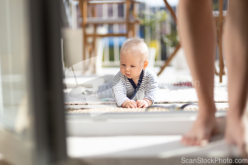 Image of Cute little infant baby boy playing with toys outdoors at the patio in summer being supervised by her mother seen in the background.