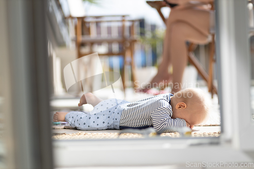Image of Cute little infant baby boy sulking while playing with toys outdoors at the patio in summer being supervised by her mother seen in the background.