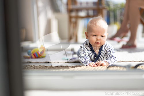 Image of Cute little infant baby boy playing with toys outdoors at the patio in summer being supervised by her mother seen in the background.