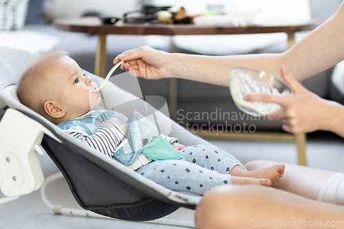 Image of Mother spoon feeding her baby boy infant child in baby chair with fruit puree. Baby solid food introduction concept.