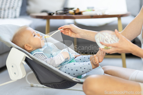 Image of Mother spoon feeding her baby boy infant child in baby chair with fruit puree. Baby solid food introduction concept.