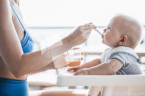 Image of Mother spoon feeding her baby boy child in baby chair with fruit puree on a porch on summer vacations. Baby solid food introduction concept.