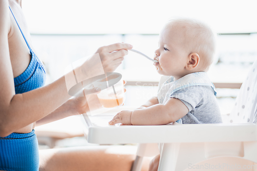 Image of Mother spoon feeding her baby boy child in baby chair with fruit puree on a porch on summer vacations. Baby solid food introduction concept.