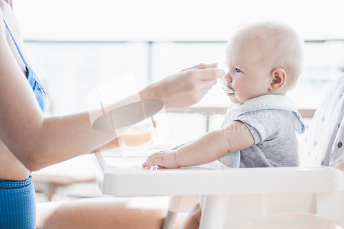 Image of Mother spoon feeding her baby boy child in baby chair with fruit puree on a porch on summer vacations. Baby solid food introduction concept.