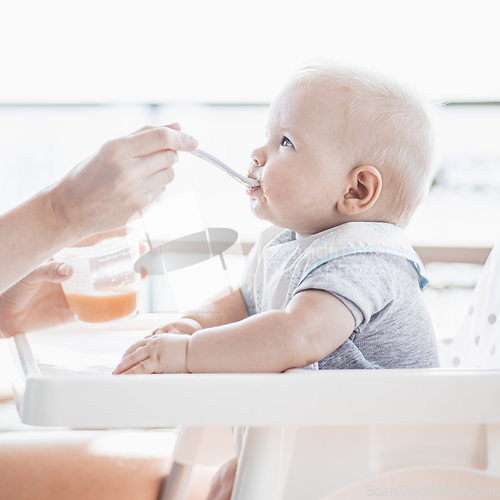 Image of Mother spoon feeding her baby boy child in baby chair with fruit puree on a porch on summer vacations. Baby solid food introduction concept.