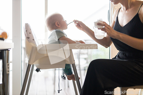 Image of Mother spoon feeding her baby boy child in baby chair with fruit puree in kitchen at home. Baby solid food introduction concept.