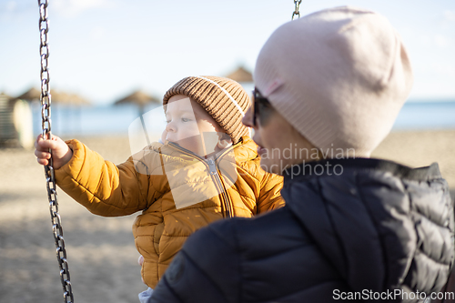 Image of Mother pushing her infant baby boy child on a swing on sandy beach playground outdoors on nice sunny cold winter day in Malaga, Spain.