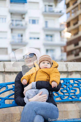 Image of Young mother with her cute infant baby boy child on bench in city park.