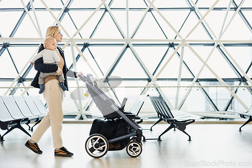 Image of Mother carying his infant baby boy child, pushing stroller at airport departure terminal moving to boarding gates to board an airplane. Family travel with baby concept.