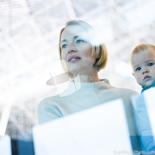 Image of Thoughtful young mother looking trough window holding his infant baby boy child while waiting to board an airplane at airport terminal departure gates. Travel with baby concept.