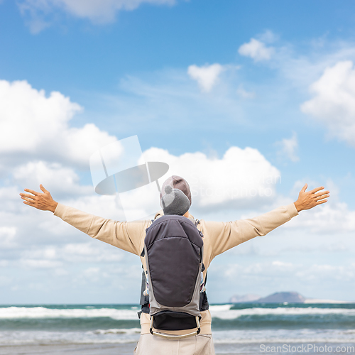 Image of Young father rising hands to the sky while enjoying pure nature carrying his infant baby boy son in backpack on windy sandy beach of Famara, Lanzarote island, Spain. Family travel concept.