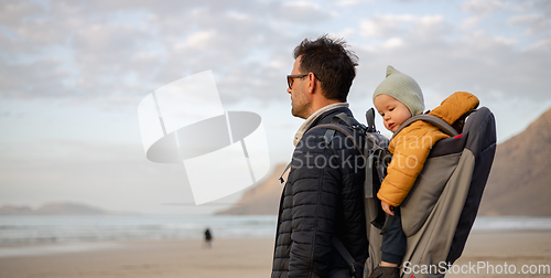 Image of Father enjoying pure nature carrying his infant baby boy son in backpack on windy sandy beach of Famara, Lanzarote island, Spain at sunset. Family travel concept.