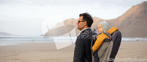 Image of Father enjoying pure nature carrying his infant baby boy son in backpack on windy sandy beach of Famara, Lanzarote island, Spain at sunset. Family travel concept.