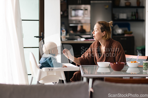 Image of Cheerful mother wearing bathrope spoon feeding her infant baby boy child sitting in high chair at the dining table in kitchen at home in the morning.