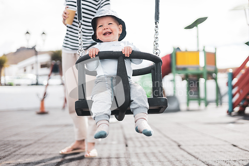 Image of Mother pushing her infant baby boy child on a swing on playground outdoors.