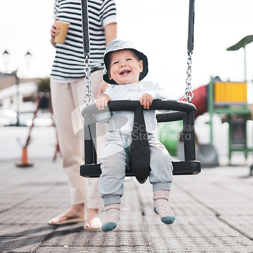 Image of Mother pushing her infant baby boy child on a swing on playground outdoors.