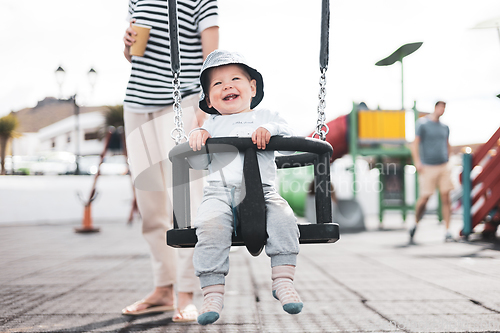 Image of Mother pushing her infant baby boy child on a swing on playground outdoors.