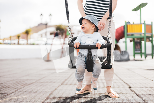 Image of Mother pushing her infant baby boy child on a swing on playground outdoors.