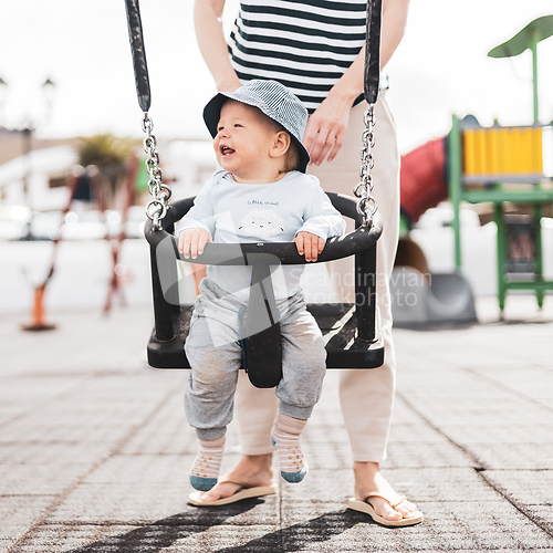 Image of Mother pushing her infant baby boy child on a swing on playground outdoors.