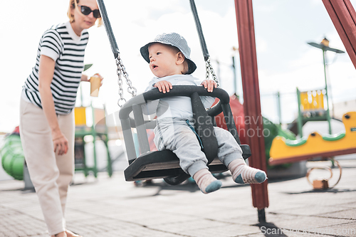 Image of Mother pushing her infant baby boy child on a swing on playground outdoors.