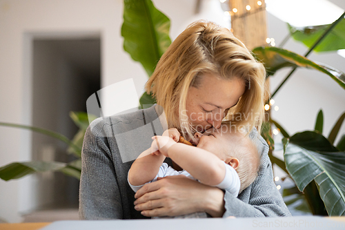 Image of Portrait of young mother cuddling and kissing her adorable little child while sitting at the table at home. Sensory stimulation for baby development