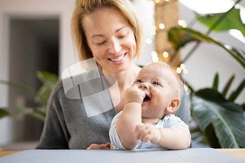 Image of Portrait of young mother cuddling her adorable little child while sitting at the table at home. Sensory stimulation for baby development