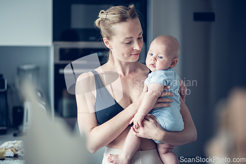 Image of Pretty young mother holding her newborn baby boy standing near kitchen window at home