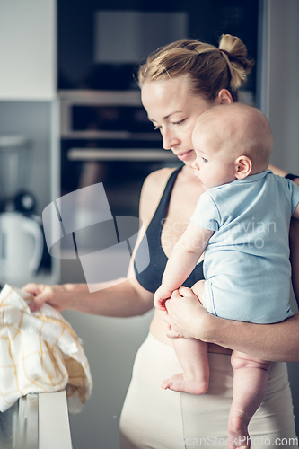 Image of Woman wiping kitchen sink with a cloth after finishing washing the dishes while holding four months old baby boy in her hands