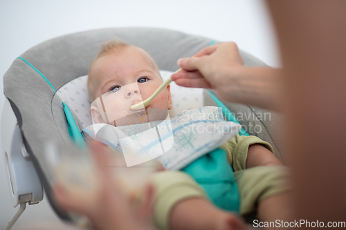 Image of Mother spoon feeding her baby boy infant child in baby chair with fruit puree. Baby solid food introduction concept.