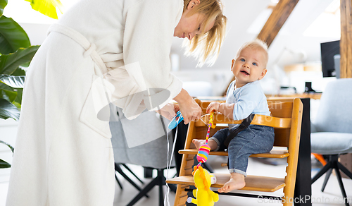 Image of Happy infant sitting and playing with his toy in traditional scandinavian designer wooden high chair in modern bright atic home suppervised by his mother.