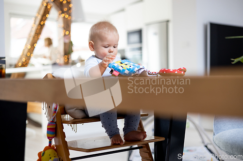 Image of Happy infant sitting at dining table and playing with his toy in traditional scandinavian designer wooden high chair in modern bright atic home. Cute baby playing with toys