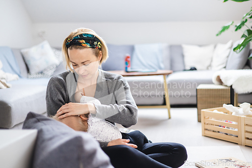 Image of Young woman breastfeeding her infant baby boy casualy sitting on child's playing mat on living room floor at home.