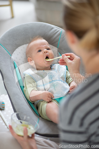 Image of Mother spoon feeding her baby boy infant child in baby chair with fruit puree. Baby solid food introduction concept.