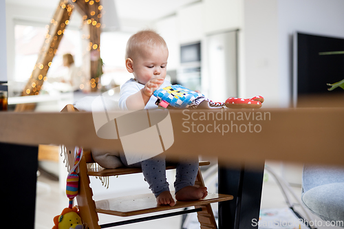 Image of Happy infant sitting at dining table and playing with his toy in traditional scandinavian designer wooden high chair in modern bright atic home. Cute baby playing with toys