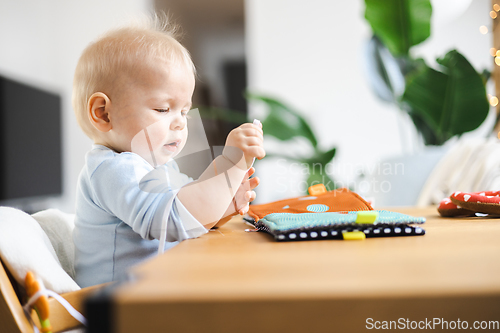 Image of Happy infant sitting at dining table and playing with his toy in traditional scandinavian designer wooden high chair in modern bright atic home. Cute baby playing with toys