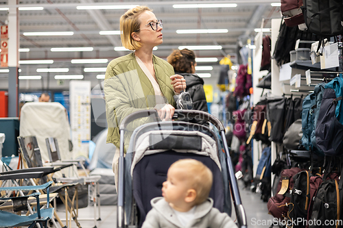 Image of Casualy dressed mother choosing sporty shoes and clothes products in sports department of supermarket store with her infant baby boy child in stroller.