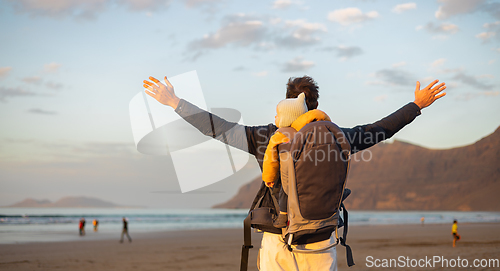 Image of Young father rising hands to the sky while enjoying pure nature carrying his infant baby boy son in backpack on windy sandy beach of Famara, Lanzarote island, Spain at sunset. Family travel concept.