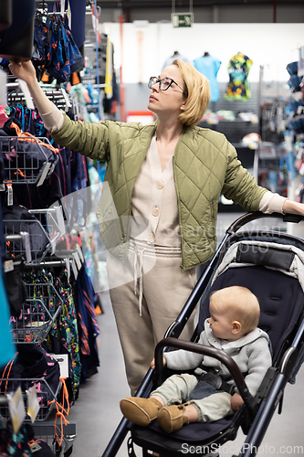 Image of Casualy dressed mother choosing sporty shoes and clothes products in sports department of supermarket store with her infant baby boy child in stroller.