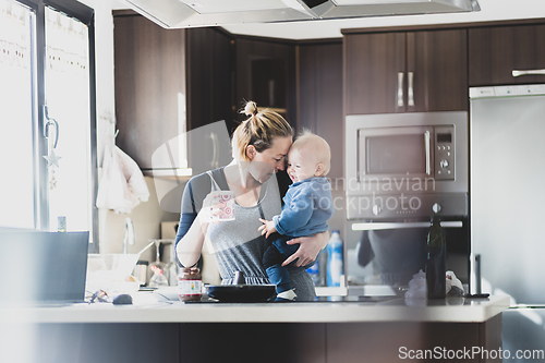 Image of Happy mother holding her little infant baby boy while drinking morning coffee and making pancakes for breakfast in domestic kitchen. Family lifestyle, domestic life concept.