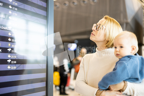 Image of Mother traveling with child, holding his infant baby boy at airport terminal, checking flight schedule, waiting to board a plane. Travel with kids concept.