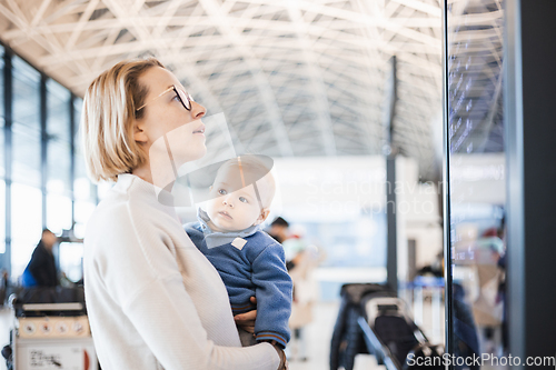 Image of Mother traveling with child, holding his infant baby boy at airport terminal, checking flight schedule, waiting to board a plane. Travel with kids concept.