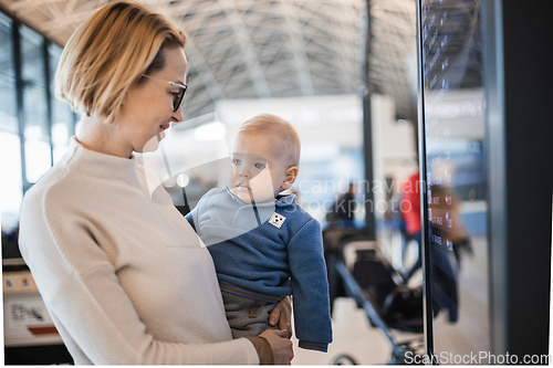 Image of Mother traveling with child, holding his infant baby boy at airport terminal, checking flight schedule, waiting to board a plane. Travel with kids concept.